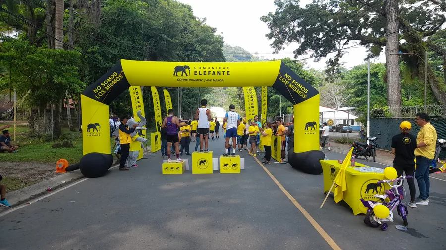 Meta de carrera de Comunidad Elefante con arco inflable amarillo y negro, banderolas, funda de mesa y podio, con corredores y público